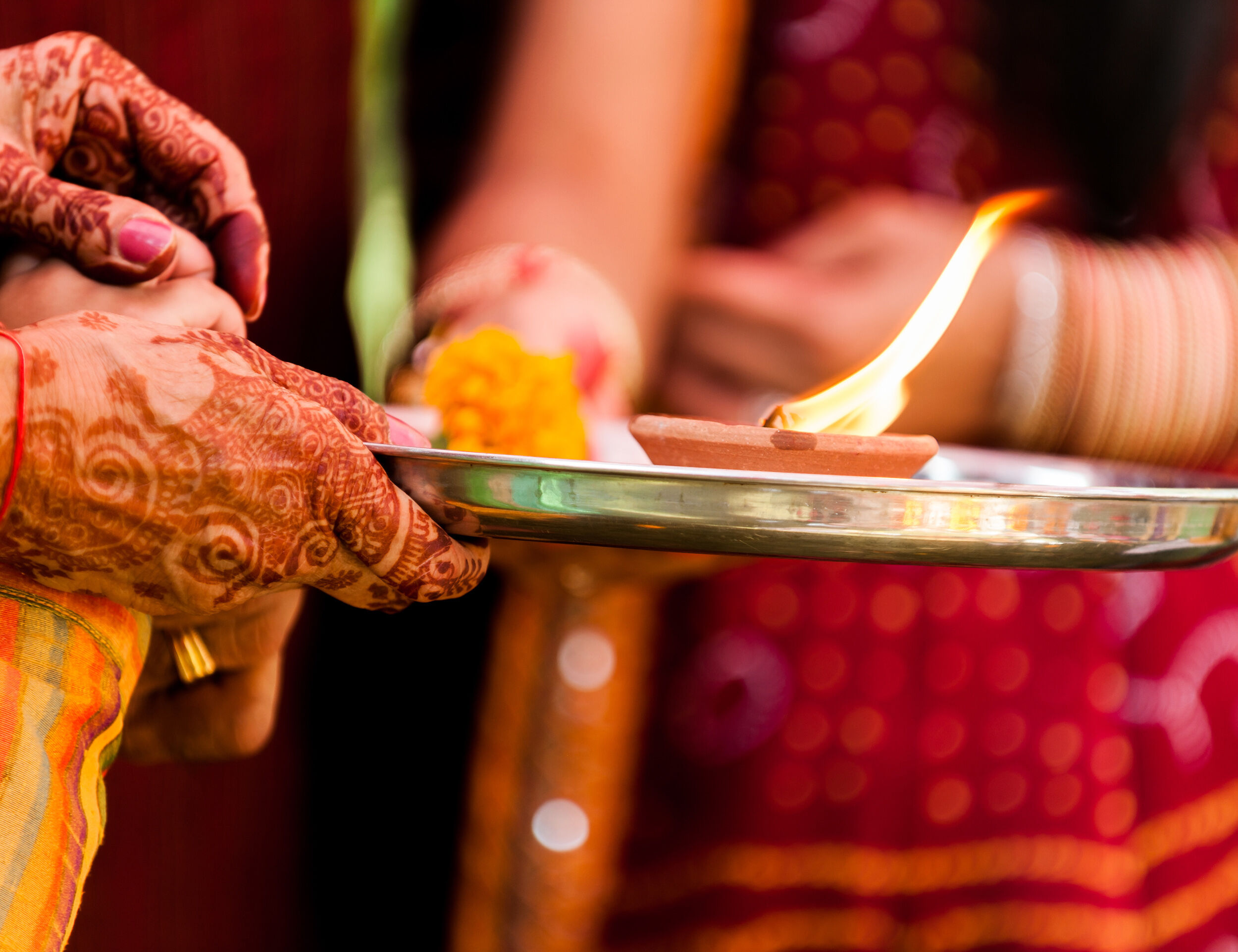 Close-up of a traditional Indian wedding ritual with hands adorned in mehndi holding a metal plate with a lit oil lamp, flowers, and ceremonial items during aarti.